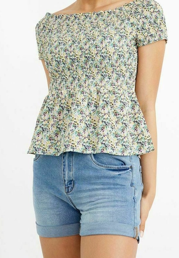 Femme portant un haut smocké à manches courtes fleuri, assorti à un short en jean bleu clair avec ourlet roulé, sur un fond uni.