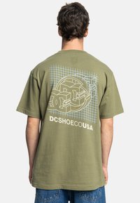 Olive green cotton t-shirt featuring a graphic design with circular shapes and grid pattern on the back. Short sleeves, relaxed fit.