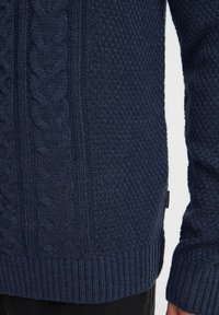 Pull en maille bleu marine avec des motifs texturés, des poignets et un ourlet côtelés, et un accent en tricot torsadé sur le côté.