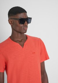 T-shirt rosso a V con una finitura testurizzata e un piccolo logo sul lato sinistro del petto; abbinato a occhiali da sole neri oversize.