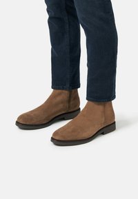 Bottines Chelsea en daim marron avec un bout rond et une semelle en caoutchouc noir, associées à un jean en denim bleu foncé, mettant en valeur une texture lisse et douce.