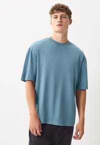 Világoskék rövid ujjú póló puha anyagból, kerek nyakkivágással és laza szabással. Sötét farmerral viselve.