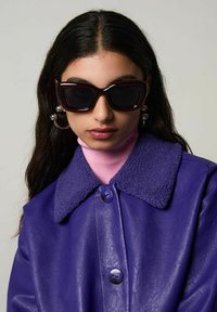 Lila läderjacka med fårskinnskrage, tillsammans med en rosa polotröja och mörka oversize-solglasögon. Utrustad med stora silverringar som örhängen.