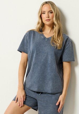 Blondynka w wyblakłej, niebieskiej, oversize'owej koszulce z dekoltem w serek i dopasowanych szortach, stojąca na tle gładkiej, białej ściany.