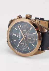 Montre-bracelet chronographe dorée avec un cadran noir, trois sous-cadrans, index beiges et un bracelet en cuir noir sur fond blanc.