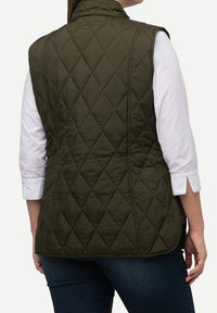 Frau, die eine dunkelgrüne gesteppte Weste über einem weißen Langarmshirt und blauen Jeans trägt, von hinten vor einem einfarbigen Hintergrund gezeigt.