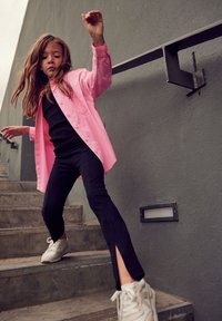 Camisa de botones rosa sobre una camiseta negra, pantalones negros acampanados con aberturas, y zapatillas blancas. Persona en movimiento sobre escalones de concreto.