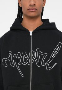 Čierna mikina na zips vyrobená z jemného materiálu, na prednej strane s veľkým bielym, stylizovaným logom "ripcurl" a s kapucňou na šnúrku.