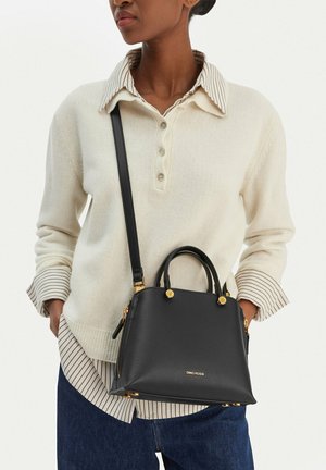 Donna che indossa un maglione crema sopra una camicia a righe e jeans blu, con una borsa nera in pelle Gino Rossi con dettagli dorati a tracolla.