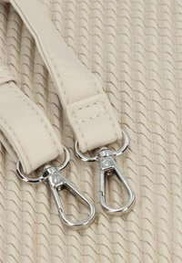 Des sangles en cuir beige clair avec des crochets à clip en métal argenté, reposant sur un fond texturé de couleur crème avec des motifs ondulés.