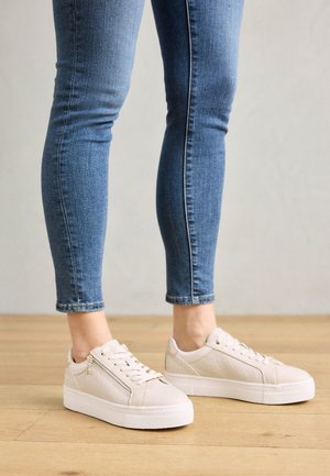 Beige sneakers with textured accents, featuring a lace-up front and a chunky white sole, paired with fitted blue jeans.