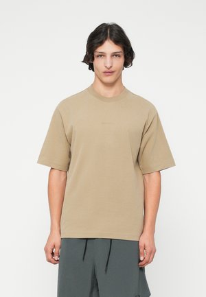 Beige oversized t-shirt with short sleeves, round neck, made of soft cotton, featuring subtle branding on the chest. Worn with gray shorts.