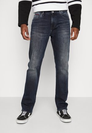 Homme portant un jean bleu foncé délavé, un t-shirt à manches longues rayé noir et blanc, et des baskets noires avec des lacets blancs, debout sur un sol gris.