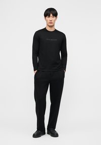 Mann steht mit Händen in den Taschen, trägt ein schwarzes Emporio Armani Langarmshirt, schwarze Hosen und schwarze Schuhe vor weißem Hintergrund.