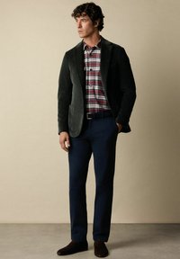 Tmavě zelený kordový blazer přes červenou kostkovanou košili, doplněný námořnickými kalhotami a hnědými botami. Jednoduchý, strukturovaný design s texturovanou tkaninou.