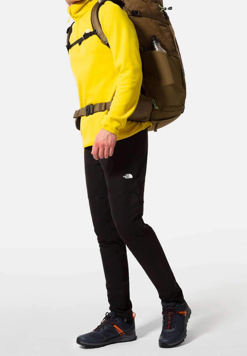 Personne portant une polaire jaune, un pantalon d'extérieur noir, des chaussures de randonnée et portant un grand sac à dos marron avec une bouteille d'eau dans la poche latérale.