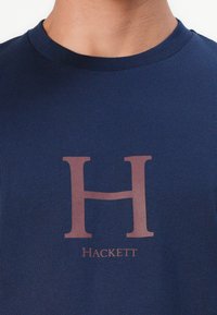 Modrá bavlnená tričko s veľkým hnedým logom "H" a slovom "Hackett" pod ním. Klasický okrúhly výstrih a hladká textúra.