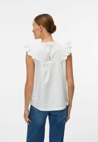 Haut en coton blanc avec des manches en dentelle froncées. Dos froncé avec une ouverture en trou de serrure. Associé à un jean large bleu.