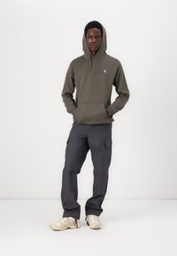 Felpa con cappuccio verde oliva con tasca frontale e logo blu, abbinata a pantaloni cargo grigi scuri con tasche laterali e sneakers chiare.