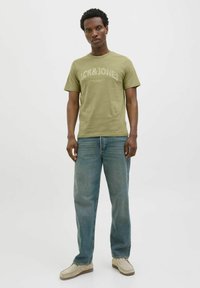 Olijfgroene katoenen T-shirt met de tekstafdruk "JACK&JONES", gecombineerd met lichtblauwe denim jeans. Model staat voor een neutrale achtergrond.