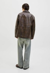Brown leather jacket with a boxy design, noticeable stitching, and a smooth texture, paired with loose light blue jeans and black shoes.