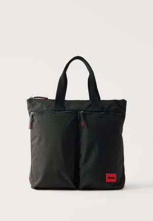Čierna kabelka tote s dvoma prednými vreckami na zips, krátkymi rukoväťami a malou červenou nášivkou "HUGO" v pravom dolnom rohu vpredu.
