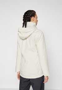 Giacca impermeabile beige chiaro con cappuccio aderente, maniche lunghe e orlo posteriore leggermente più lungo. Tessuto liscio e dettagli di design minimali.