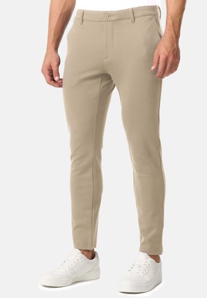 Mann trägt enge beige Hose und weiße Sneakers, steht vor schlichtem Hintergrund, Hände entspannt an den Seiten.