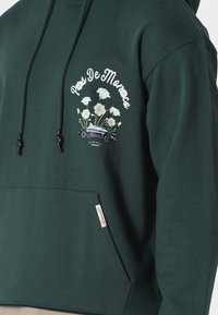 Felpa con cappuccio verde scuro con motivo floreale bianco, grafica di un'auto d'epoca e testo "Pas De Monaco" sopra la tasca sul petto.