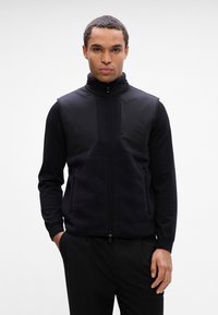 Colete preto com fecho éclair e gola alta, textura de polar na parte inferior e tecido liso na parte superior. Apresenta dois bolsos laterais e detalhes com o logótipo.