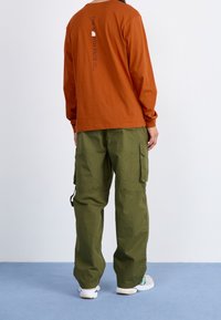 Koszula z długim rękawem w kolorze rdzy z graficznym nadrukiem na plecach, połączona z luźnymi oliwkowymi spodniami cargo z bocznymi kieszeniami.