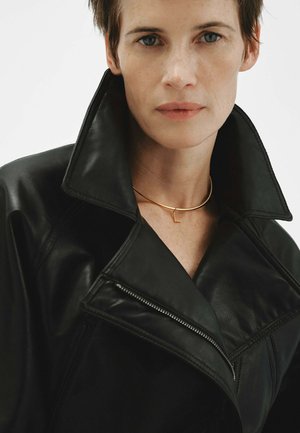 Mujer con cabello corto que lleva una chaqueta de cuero negra con cuello ancho y un collar dorado con un pequeño colgante.