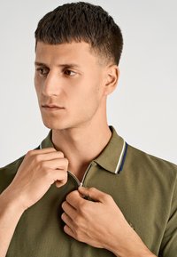 Olivgrünes Polohemd mit Reißverschluss und einem strukturierten Kragen mit blauen und weißen Streifen. Das Model zieht den Reißverschluss mit beiden Händen hoch.