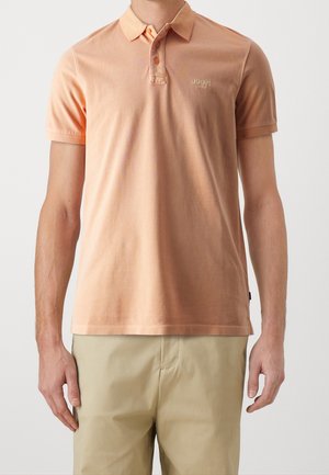 Man draagt een perzikkleurig poloshirt met korte mouwen en twee knopen, gecombineerd met beige broek, staande tegen een effen lichte achtergrond.