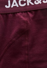 Κοντινό πλάνο βουργουνδί υφάσματος με το «JACK&» να φαίνεται στη μέση, παρουσιάζοντας λεπτομερή ραφή και υφή.