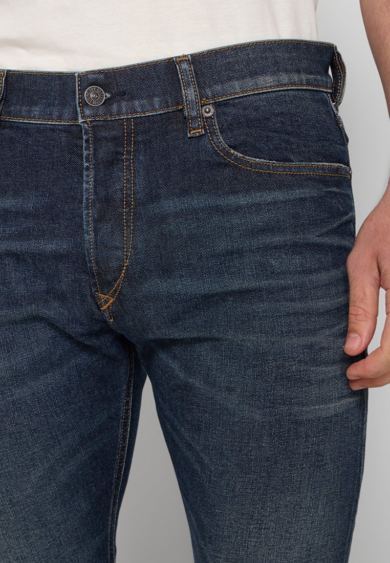 Nahaufnahme einer Person, die dunkelblaue Jeans mit orangefarbenen Nähten und ein weißes Hemd trägt, mit der Hand in der Nähe der Tasche.