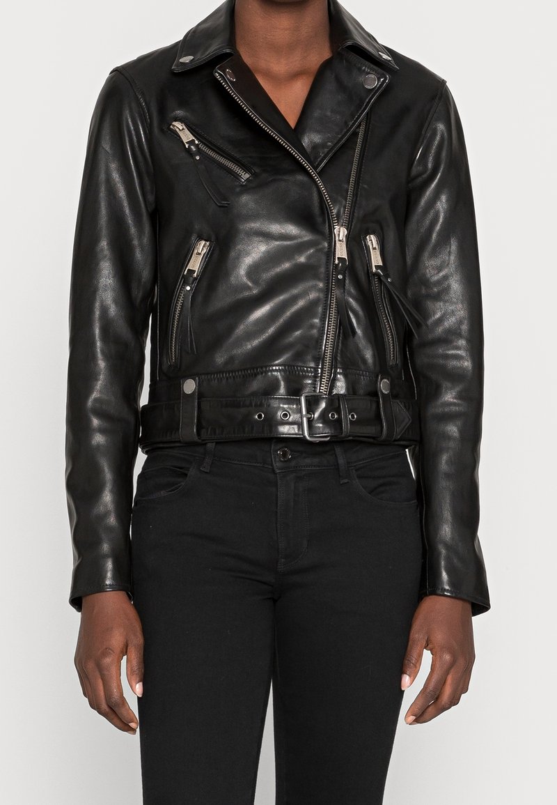 Veste de motard en cuir noir avec poches zippées, larges revers et taille ceinturée portée sur un pantalon noir sur une personne debout.