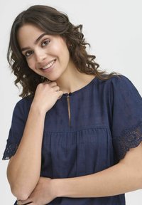 Blusa blu navy con maniche corte, decorata con pizzo, colletto rotondo e un piccolo spacco anteriore, realizzata in un tessuto leggero e texturizzato.