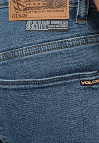 Blue denim jeans featuring a leather patch with logo and text, alongside stitching details and a tag emphasizing water conservation.