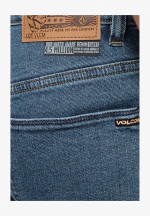 Blauwe denimjeans met een leren patch met logo en tekst, inclusief stikdetails en een label dat de nadruk legt op waterbesparing.