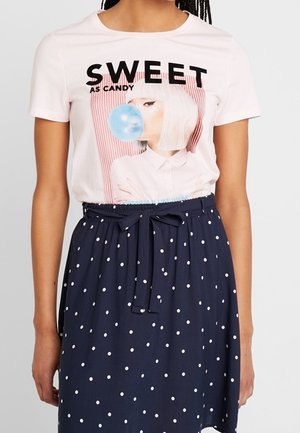 Vrouw draagt een wit T-shirt met de tekst "SWEET AS CANDY" en een afbeelding van een vrouw die een blauwe bel blaast, gecombineerd met een marineblauwe polkadot rok.