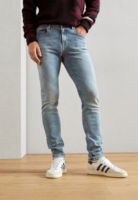 Světle modré slim-fit džíny s vybledlým umytím, v kombinaci s bílými teniskami s černými pruhy. Texturovaný svetr s vínovými akcenty.