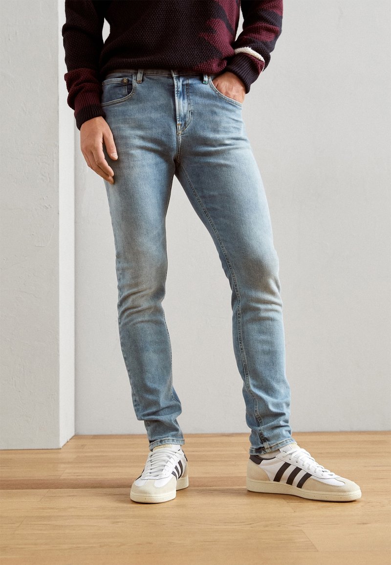 Světle modré slim-fit džíny s vybledlým umytím, v kombinaci s bílými teniskami s černými pruhy. Texturovaný svetr s vínovými akcenty.