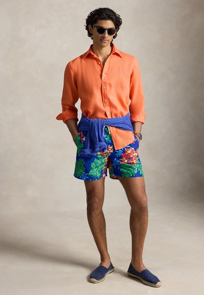 Νέος άνδρας που φοράει πορτοκαλί πουκάμισο με κουμπιά, μπλε τροπικά σορτς, μπλε πουλόβερ δεμένο στη μέση, ναυτικό espadrilles και σκούρα γυαλιά ηλίου.