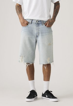 Mann trägt hellblaue, ausgewaschene Jeansshorts, weiße Socken, schwarze Turnschuhe und ein weißes T-Shirt, dabei sind tätowierte Arme sichtbar.