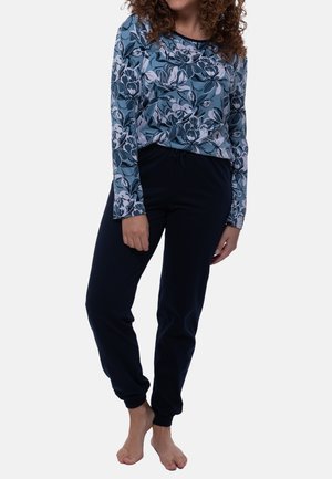Femme portant une chemise à manches longues bleue à fleurs et un pantalon de jogging noir, debout pieds nus devant un fond uni.