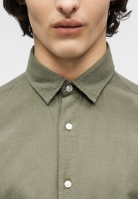 Κοντινό πλάνο ενός ατόμου που φοράει πουκάμισο ελιάς με κουμπιά και γιακά, εμφανίζονται τα χείλη, το πηγούνι και ο λαιμός.