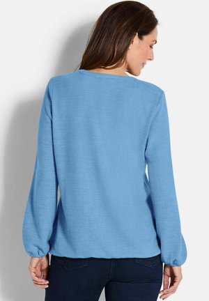 Eine Frau, die sich wegdreht, trägt einen hellblauen Langarmpullover mit gerafften Manschetten und dunklen Jeans vor einem hellen, schlichten Hintergrund.
