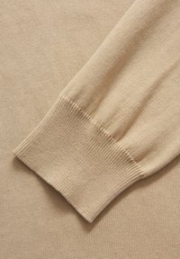 Manica di un maglione a maglia beige che mostra un polsino a coste. Il materiale è morbido, con una texture liscia e una cucitura fine e uniforme su tutta la stoffa.
