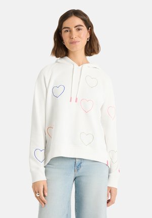 Frau trägt einen weißen Hoodie mit mehrfarbigen, gestickten Herzmustern und hellblauen Jeans, steht vor einem einfarbigen Hintergrund.